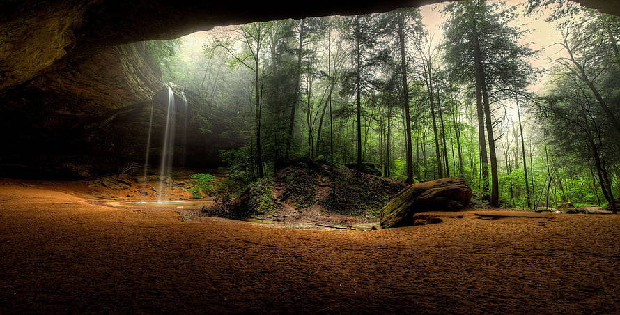 Misty forest waterfall viewed from a dark cave opening with lush green trees HD wallpaper