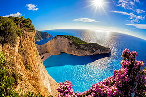 Navagio Bay in Zakynthos Greece with turquoise waters, limestone cliffs, and pink wildflowers under a bright sun HD wallpaper
