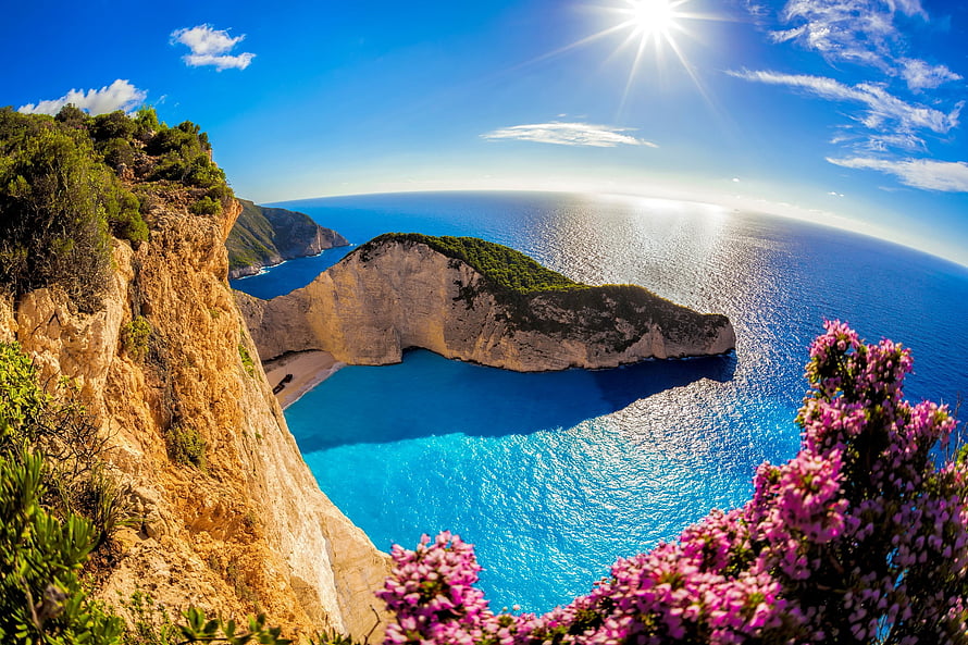 Navagio Bay in Zakynthos Greece with turquoise waters, limestone cliffs, and pink wildflowers under a bright sun HD wallpaper