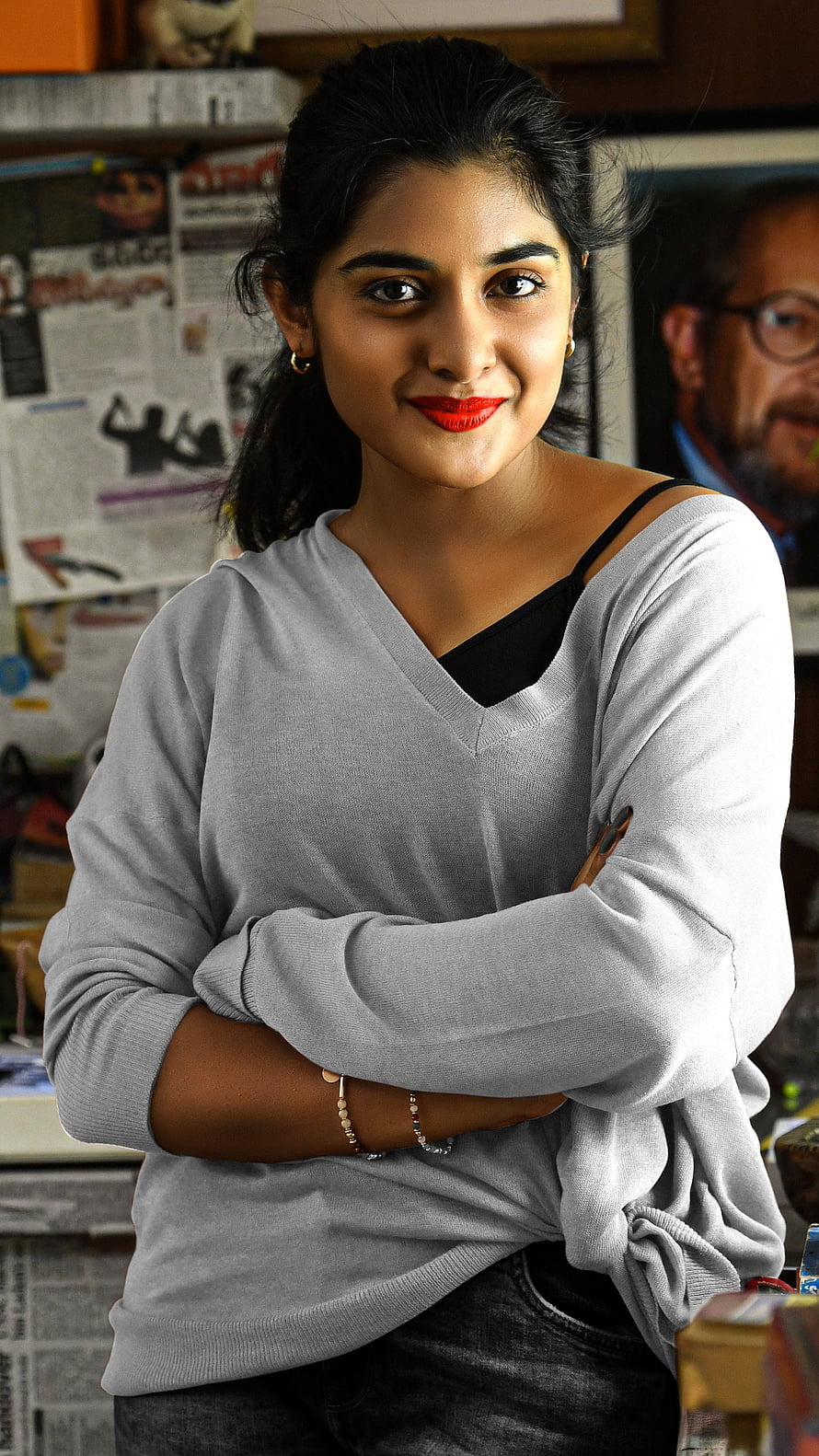 Nivetha Thomas in a grey sweater with arms crossed in a newsroom setting HD phone wallpaper