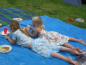 Two young girls lying on a blue picnic blanket eating outdoors on a sunny day HD wallpaper