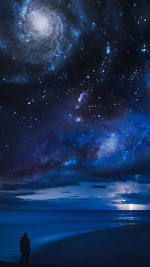 Lone figure on a dark beach beneath a vast spiral galaxy and star-filled night sky HD phone wallpaper
