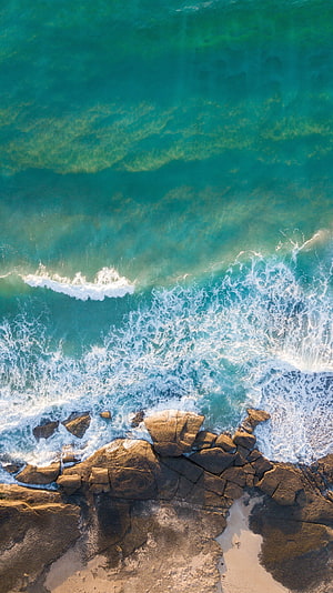 Aerial View of Turquoise Ocean Waves Crashing Against Rugged Coastal Rocks HD phone wallpaper