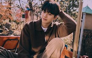 Song Kang in a brown sweater sitting outdoors with autumn foliage and birdhouses in the background HD wallpaper