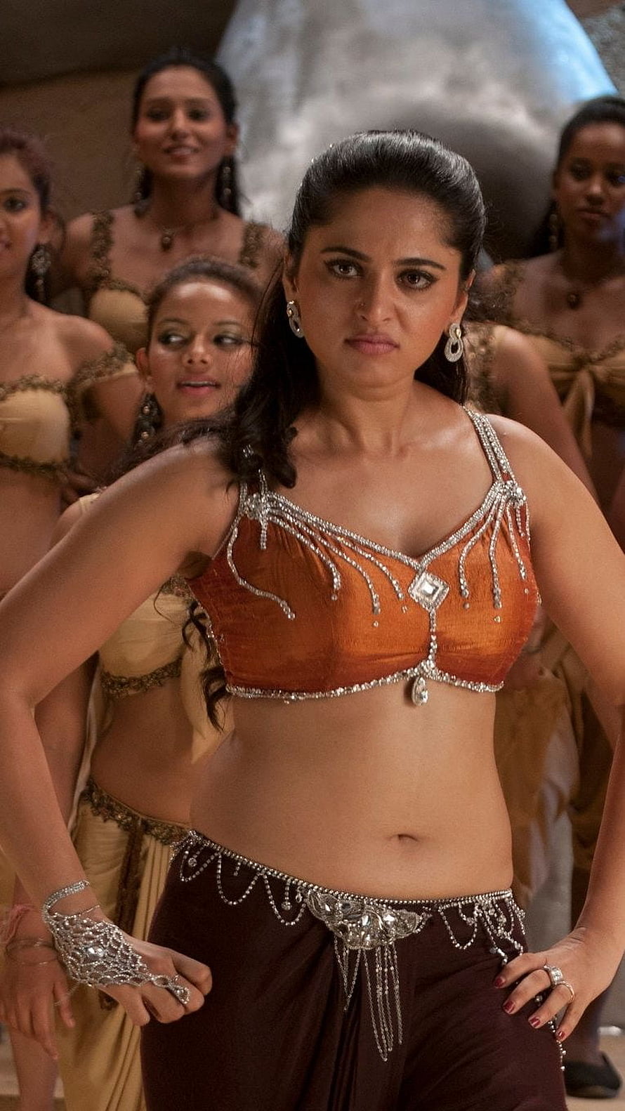 Anushka Shetty in ornate orange belly dance costume with silver jewelry and dancers behind HD phone wallpaper
