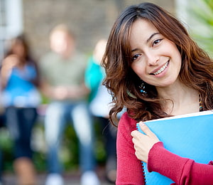 Smiling female student holding a blue notebook on a university campus HD wallpaper