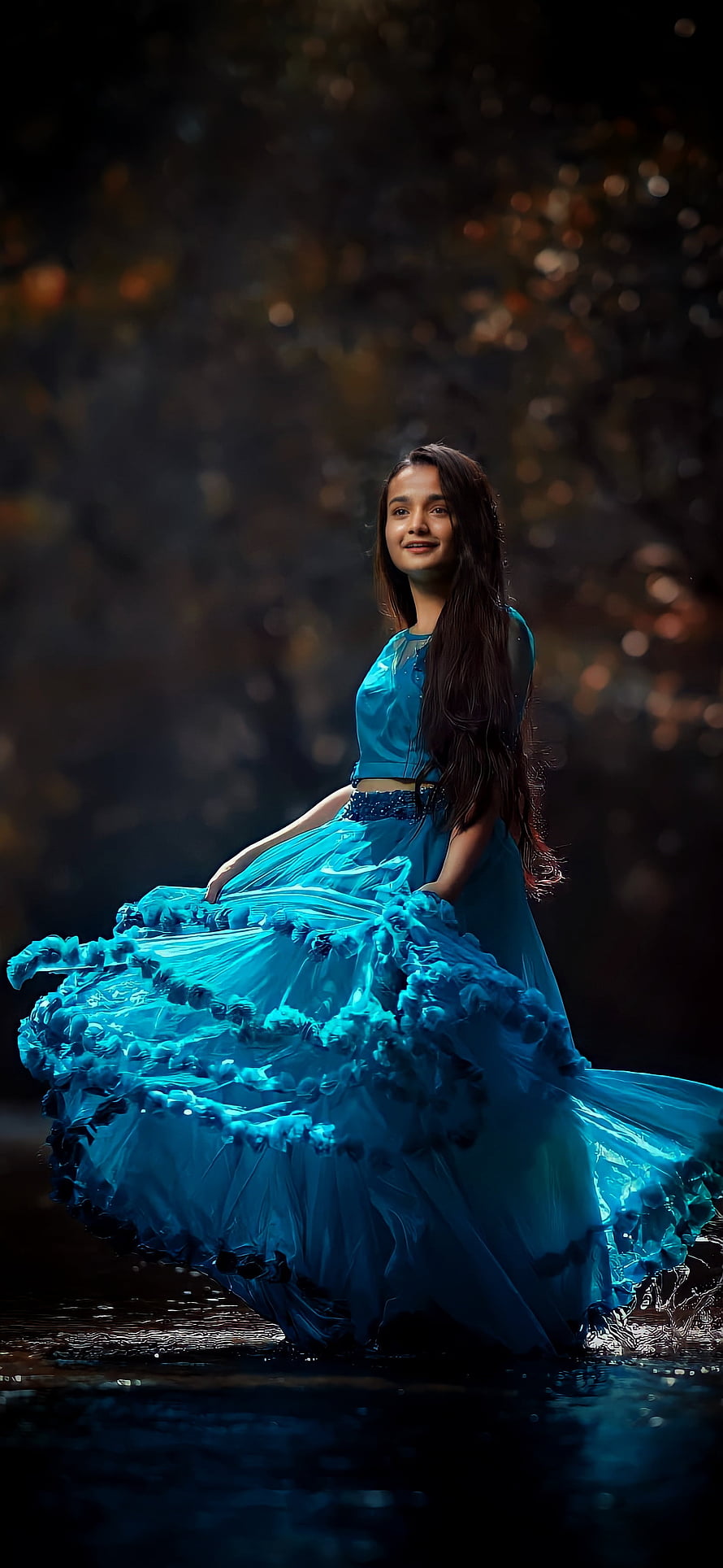 Young woman in flowing turquoise gown standing in shallow water with dark forest bokeh background HD phone wallpaper