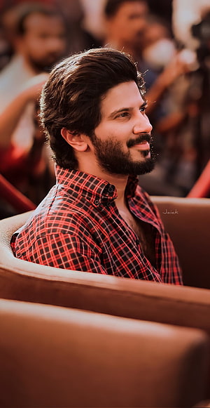 Dulquer Salmaan in a red plaid shirt with a full beard, seated in a warm-toned event setting HD phone wallpaper