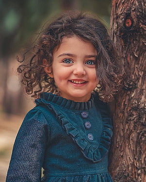 Anahita Hashemzadeh smiling portrait in a teal dress leaning against a tree trunk HD phone wallpaper