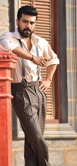Ram Charan in vintage suspenders and plaid trousers leaning on a red pillar HD phone wallpaper