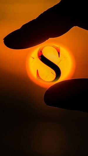 Glowing Letter S Silhouette Framed by Fingers Against a Warm Orange Sun HD phone wallpaper