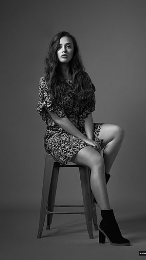 Katherine Langford in a patterned dress seated on a stool in a black and white studio portrait HD phone wallpaper