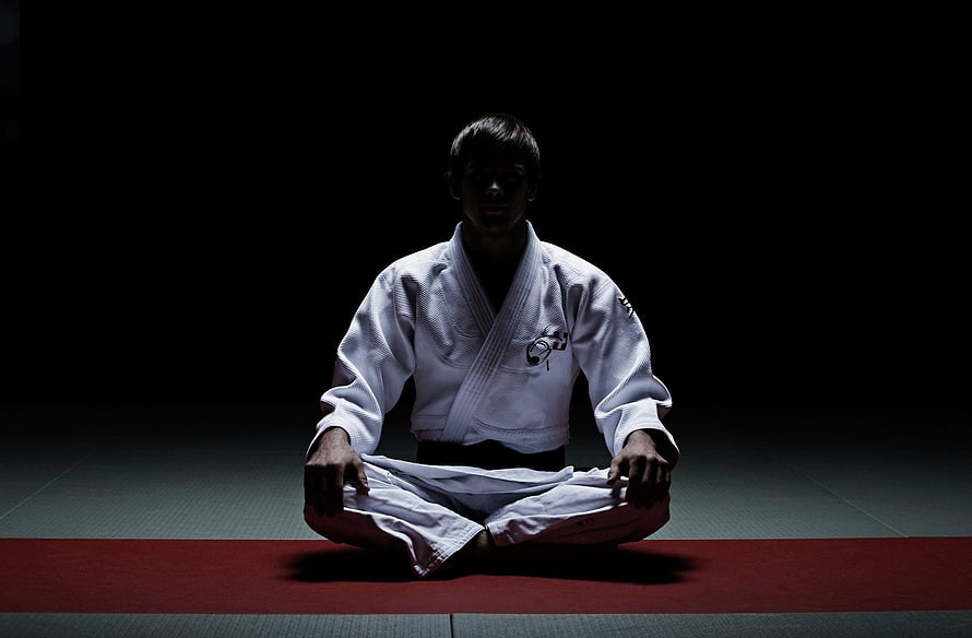 Judo practitioner in white gi meditating in seiza position on a dark dojo mat HD wallpaper