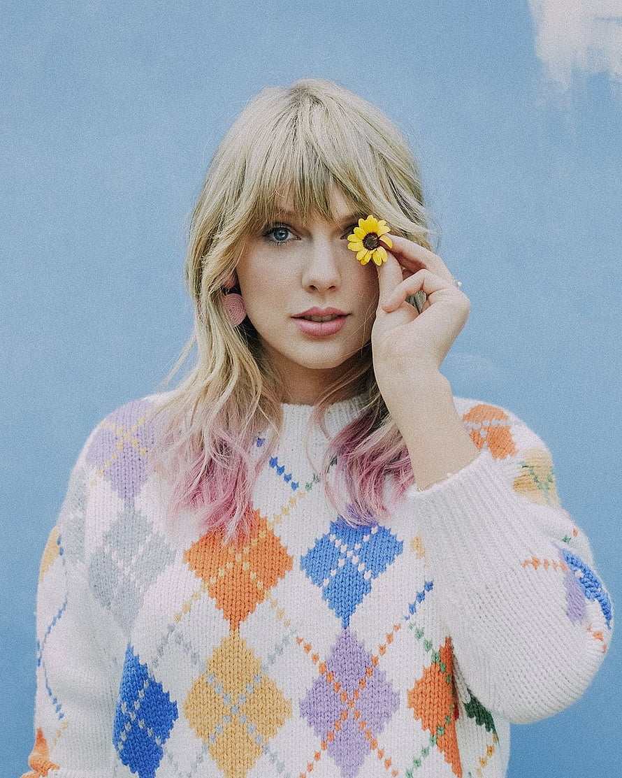 Taylor Swift in a colorful argyle sweater holding a yellow flower against a blue background HD phone wallpaper