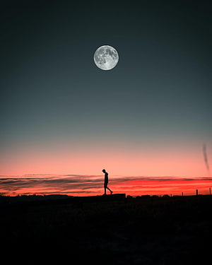 Solitary figure silhouetted against a vivid sunset sky with a large full moon overhead HD phone wallpaper