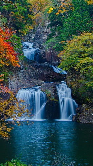 Multi-tiered waterfall cascading through a rocky gorge surrounded by vibrant autumn foliage and a deep blue pool HD phone wallpaper