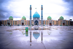 Jamkaran Mosque in Mashhad with turquoise domes and minarets reflected in a wet plaza under a cloudy sky HD wallpaper