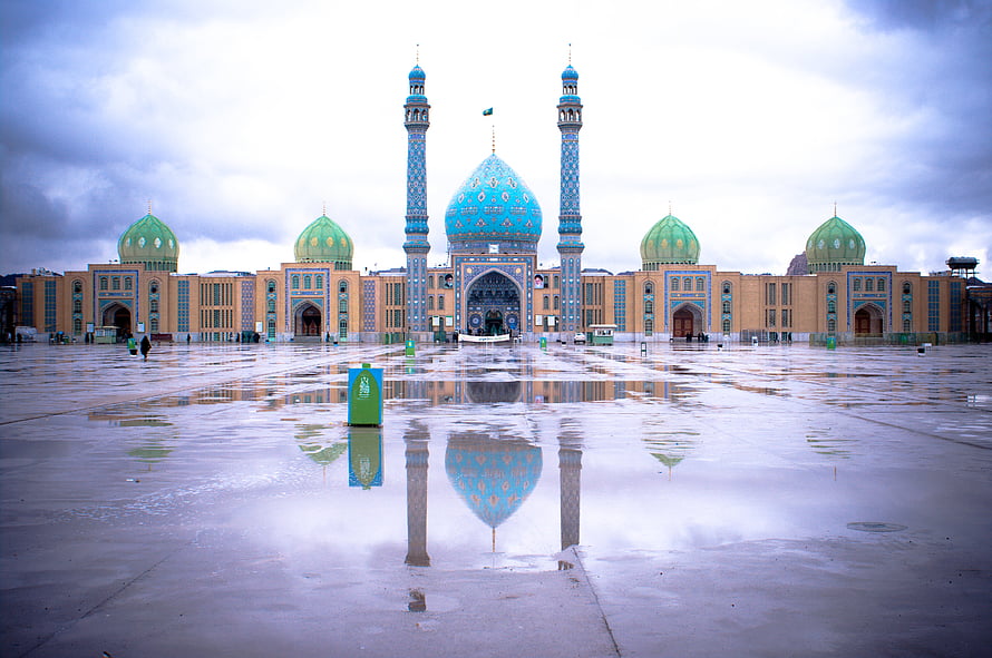 Jamkaran Mosque in Mashhad with turquoise domes and minarets reflected in a wet plaza under a cloudy sky HD wallpaper