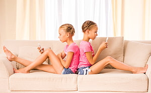 Twin girls in matching pink shirts sitting back to back on a beige sofa using smartphones HD wallpaper