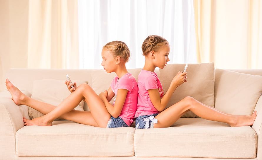 Twin girls in matching pink shirts sitting back to back on a beige sofa using smartphones HD wallpaper