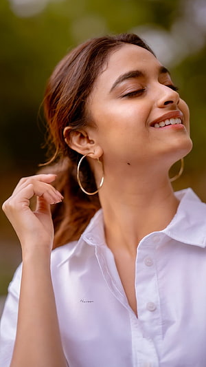 Keerthy Suresh smiling with eyes closed in a white shirt and gold hoop earrings HD phone wallpaper