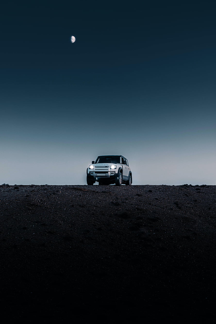 Land Rover Defender SUV on a dark desert ridge under a crescent moon at night HD phone wallpaper