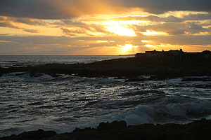Golden sunset over a rugged coastal shoreline with crashing waves and silhouetted buildings HD wallpaper