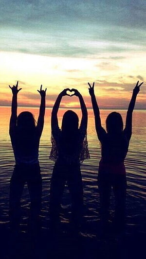 Three friends silhouetted against a vibrant sunset over the ocean making peace and heart signs HD phone wallpaper